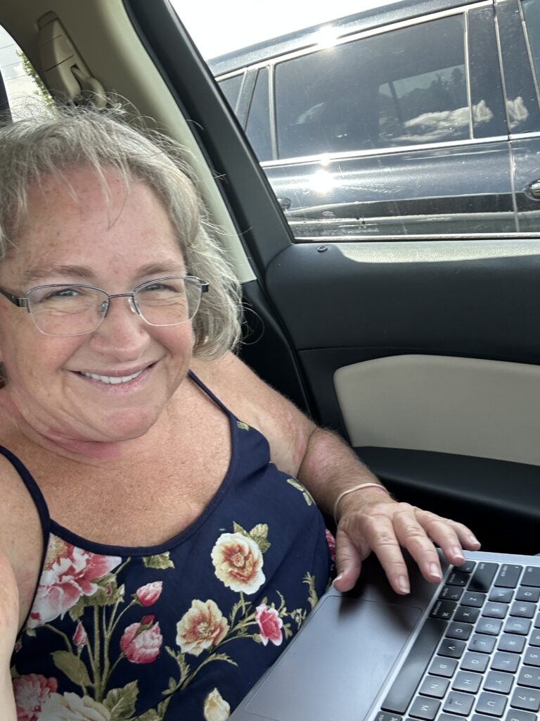 A photo of Amy smiling and working on her laptop in the passenger seat of a car. 