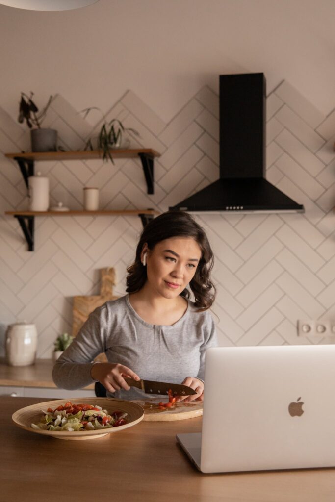 Ethnic woman preparing vegetable salad while watching laptop in kitchen